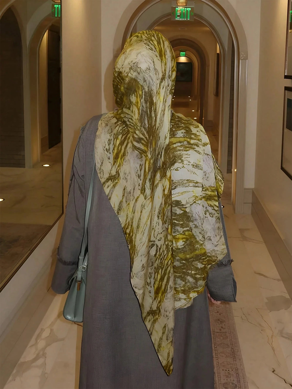 Eine Frau in einem fließenden grauen Mantel und einem eleganten gemusterten gelb-weißen Hijabschal steht in einem Flur.
