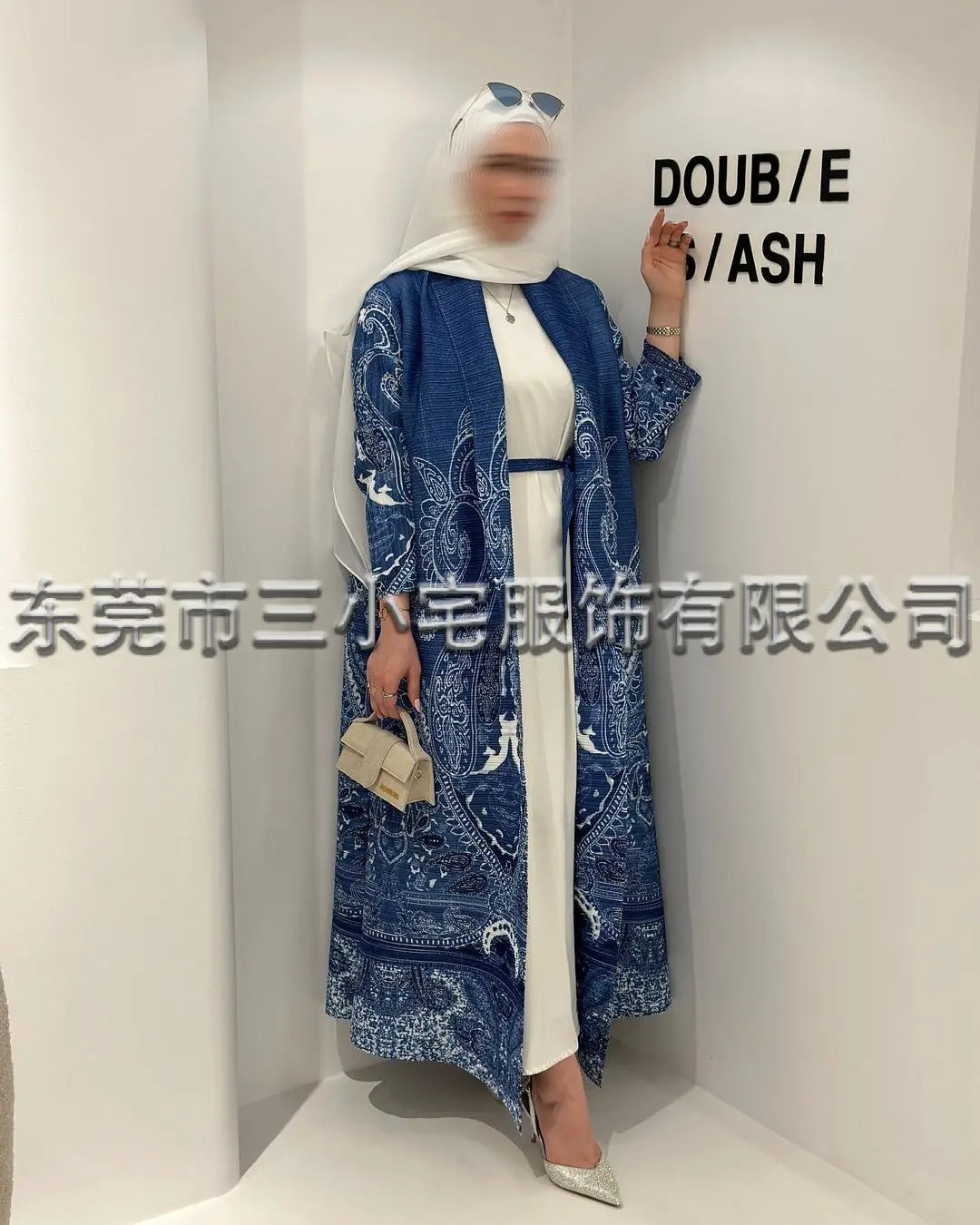 Una mujer vistiendo un cárdigan abaya de patrón azul y hijab blanco, de pie en un expositor blanco.