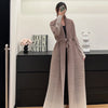 Une femme portant un trench-coat à manches longues plissé beige devant un meuble sombre.