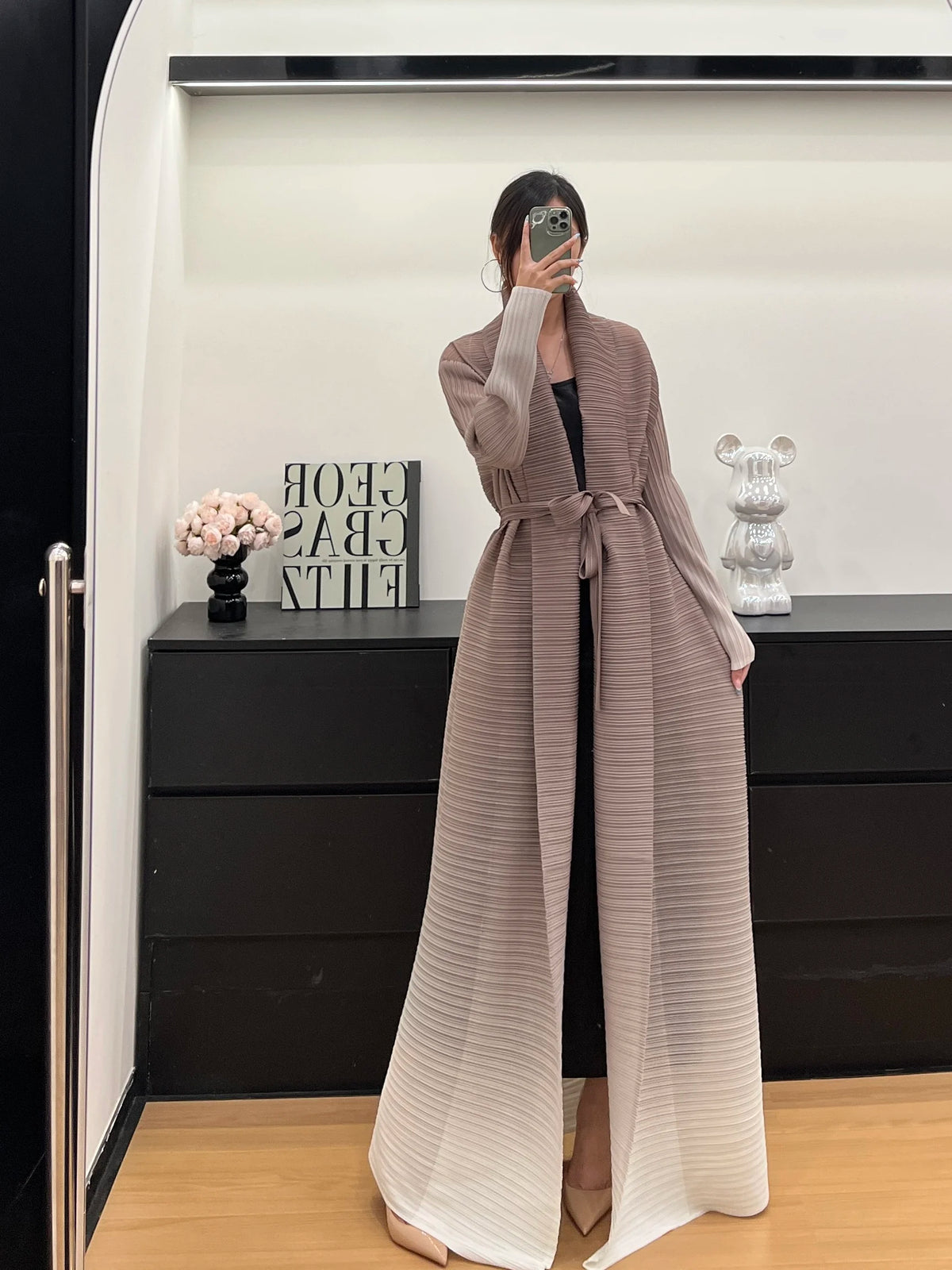 Une femme portant un trench-coat à manches longues plissé beige devant un meuble sombre.