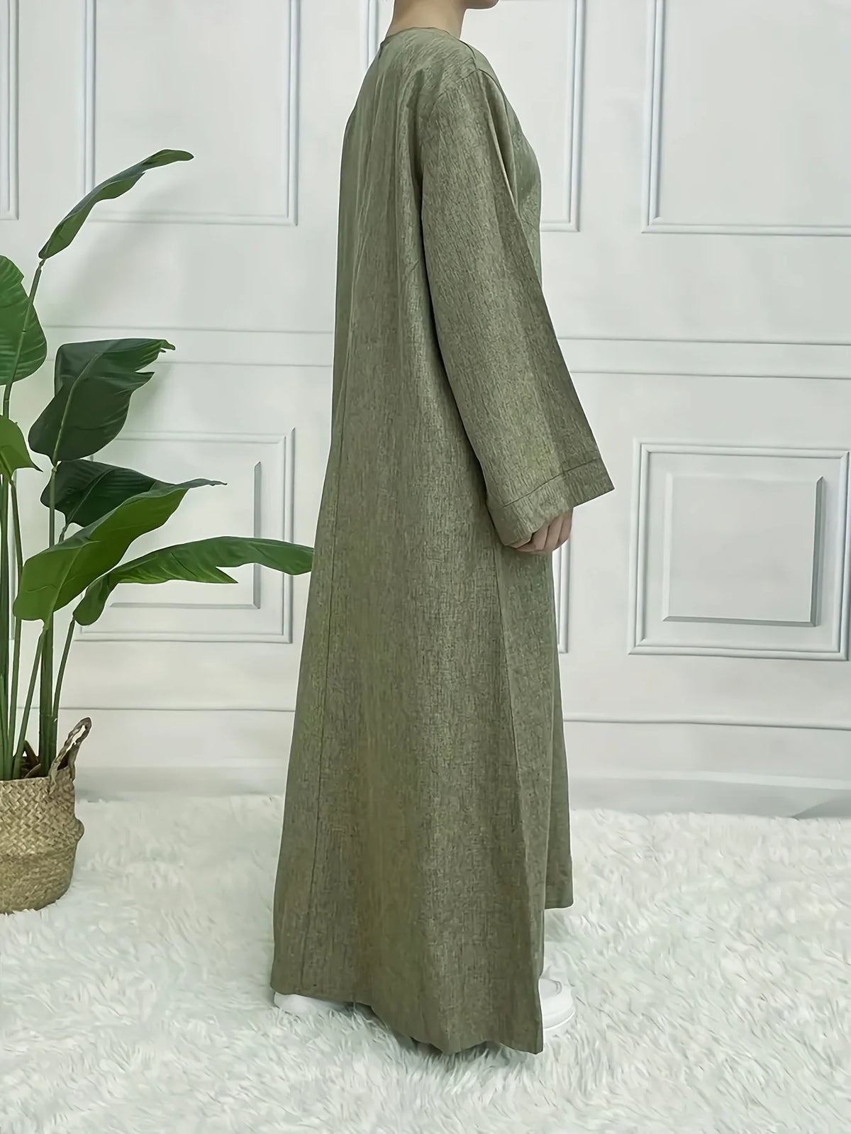 Vestido caftán holgado verde oliva completo colgado sobre una alfombra blanca peludita contra una pared de paneles blancos.
