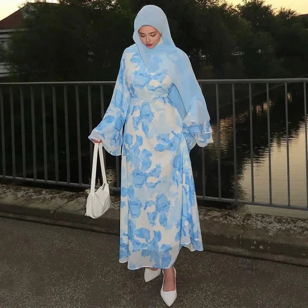 A woman wearing a light blue floral print abaya and kaftan dress with a matching hijab, standing outdoors by water.