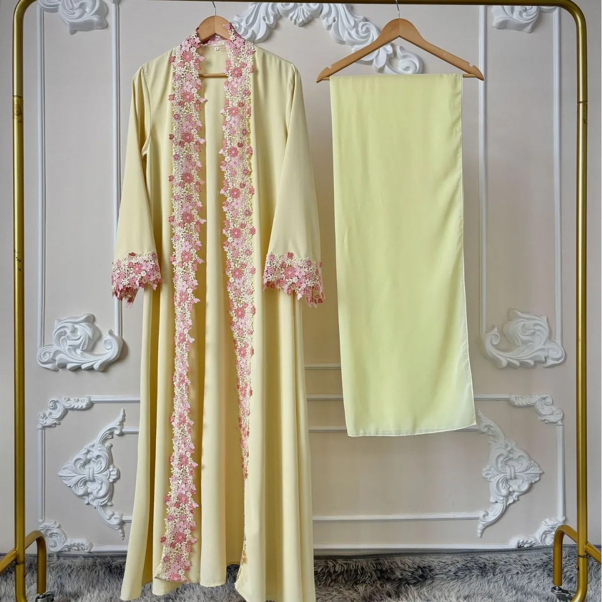 Une longue abaya jaune pâle avec des détails en dentelle florale rose, présentée sur un cintre sur fond blanc.