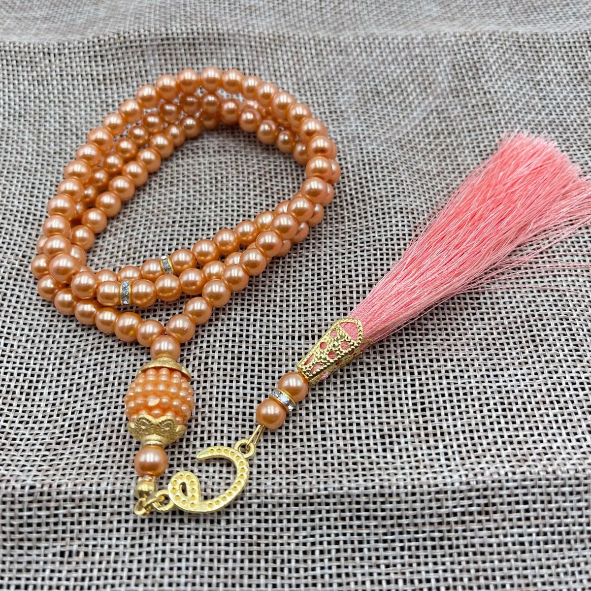 Brauner Perlenrosaryarmband mit goldfarbigem Quaste auf grauem Stoffhintergrund.