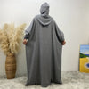 Une abaya grise à manches batwing et capuche, portée contre un mur blanc.