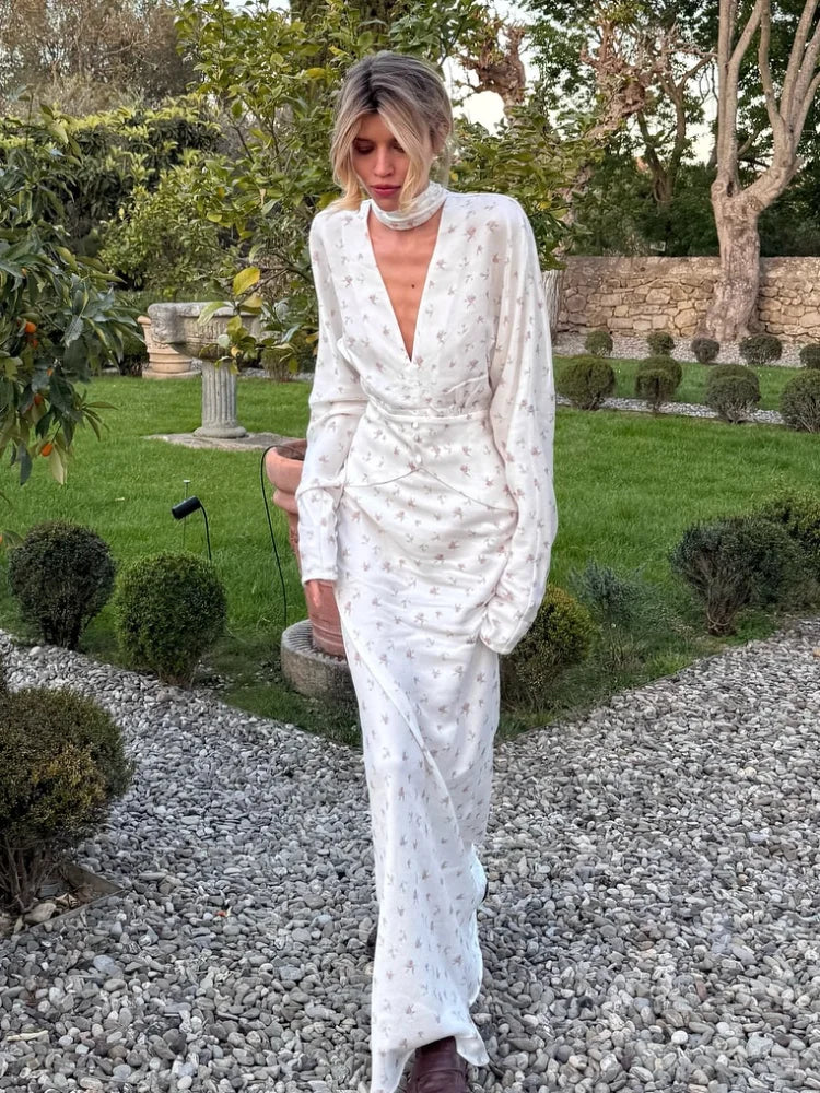 Una mujer vistiendo un vestido largo de satén con estampado floral blanco, de manga larga, de pie en un jardín.