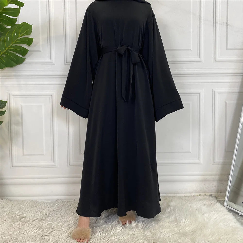 Eine elegante schwarze Maxi-Abaya für Frauen wird vor einer weißen Paneelwand präsentiert.