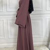 Eine Frau trägt eine elegante mauve Abaya mit schwarzen Akzenten, steht in einem gut beleuchteten Raum.