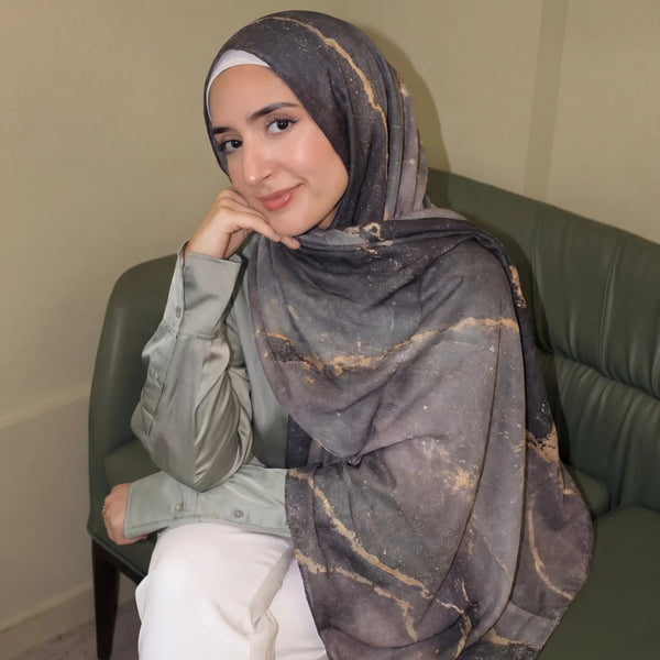 A woman wearing a gray watercolor print shawl hijab and a light green shirt sitting on a dark sofa.