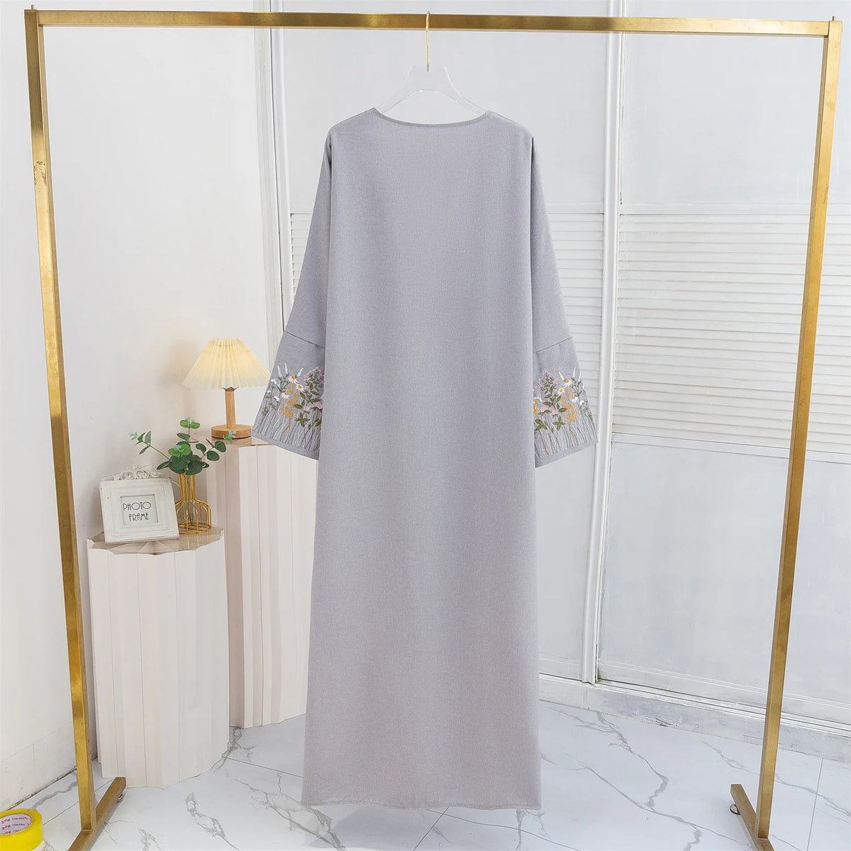 Lange, hellgraue Abaya mit floralen Stickereien in einem goldgerahmten Display.