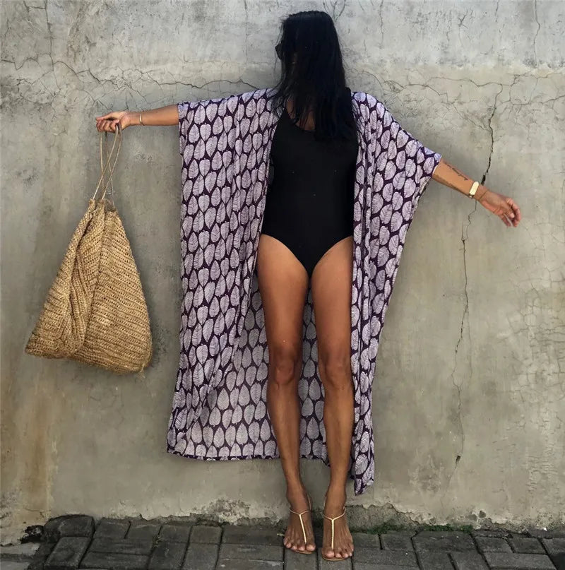 Eine Frau in einem lila gemusterten Kimono-Überwurf über einem schwarzen Kleid steht vor einer strukturierten Wand.