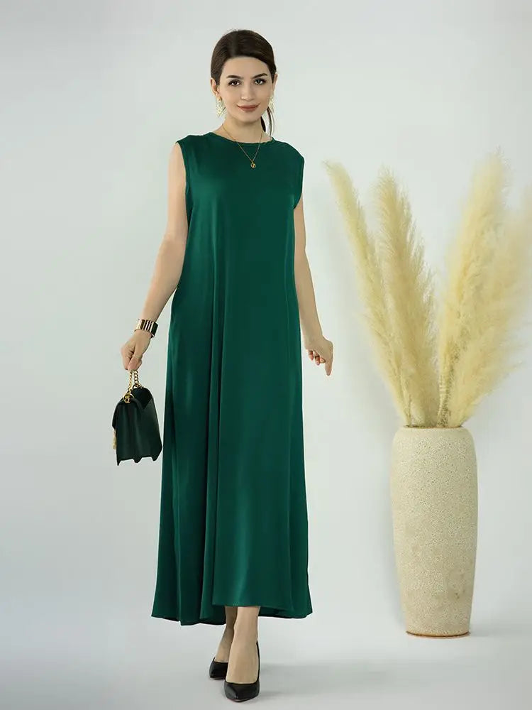 A woman models a flowing, emerald green modest abaya dress with black accessories against a white background.