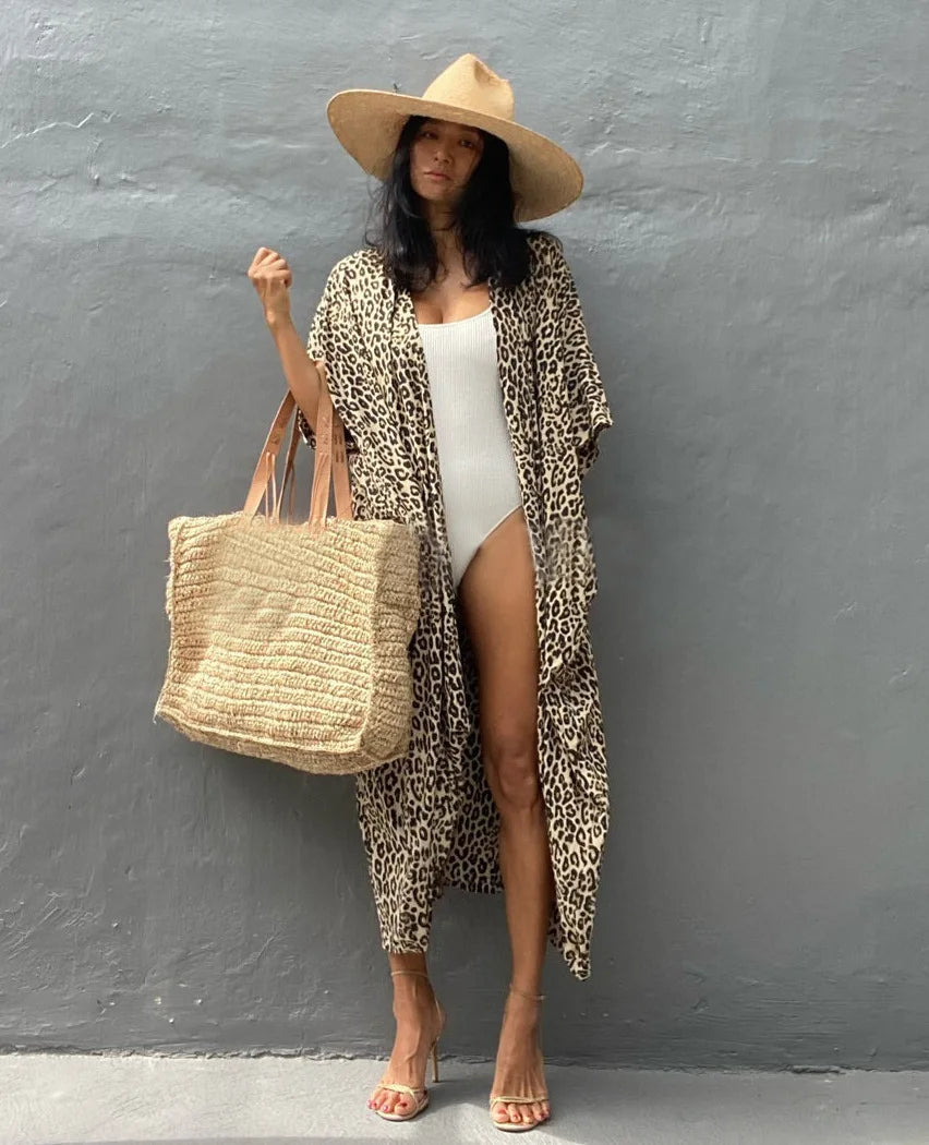 Eine Frau in leopardmuster-Kimono-Überwurf über einem weißen Mini-Kleid und einem Strohhut vor einer grauen Wand.