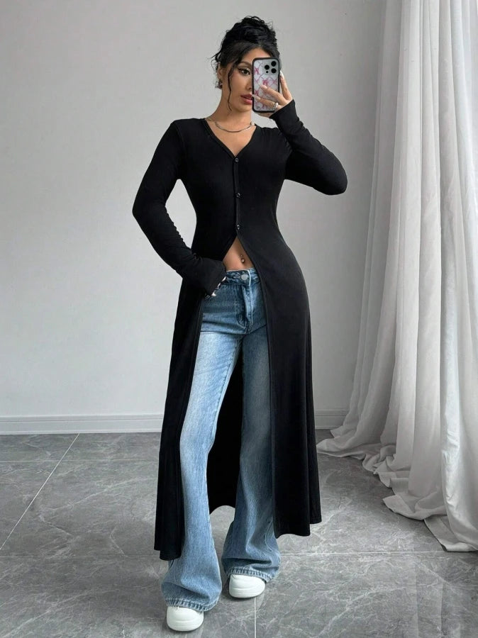 Eine Frau modelliert einen schwarzen, gerippten Strick-Maxirober über blauen Jeans, posiert drinnen.