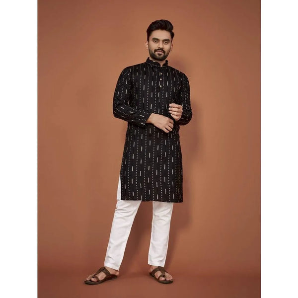 A man wearing a black patterned jubba thobe and white pants standing against a solid brown background.