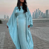 Una mujer vistiendo una abaya de satén azul claro y fluida, de corte batwing, de pie al aire libre con un horizonte de ciudad