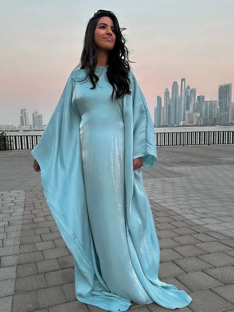 Una mujer vistiendo una abaya de satén azul claro y fluida, de corte batwing, de pie al aire libre con un horizonte de ciudad