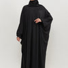 Una mujer vistiendo una elegante abaya negra con capucha y hijab, de pie contra un fondo claro.