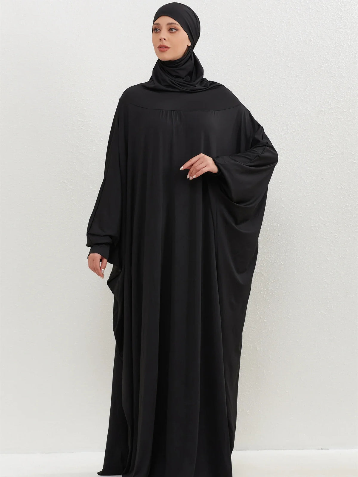 Una mujer vistiendo una elegante abaya negra con capucha y hijab, de pie contra un fondo claro.