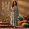 A woman models a long floral maxi dress with lantern sleeves in green and pink tones against a warm, earthy background.