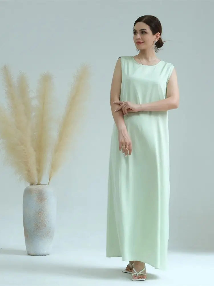 A woman wearing a light mint green flowing modest abaya dress stands next to a vase of dried ornamental grass.