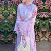 Eine Frau in einem rosa floral gemusterten Abaya und Kaftan steht vor einem grünen Hecke-Hintergrund.