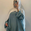 Eine Frau trägt eine leichte, fließende, bodenlange, hellblaue Abaya mit weiten Ärmeln und einen weißen Hijab und macht ein S