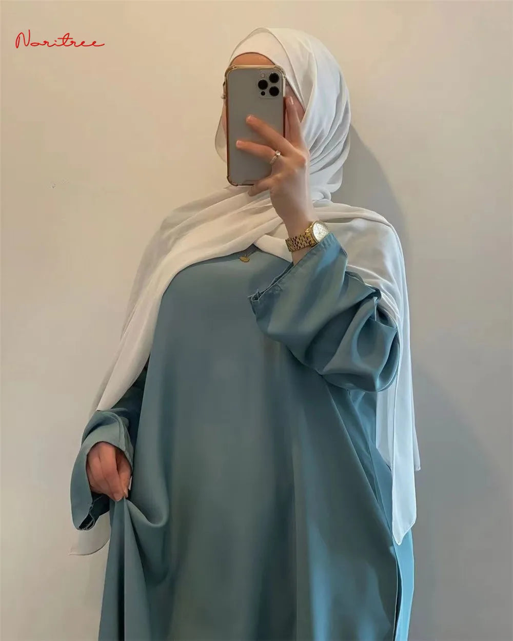 Eine Frau trägt eine leichte, fließende, bodenlange, hellblaue Abaya mit weiten Ärmeln und einen weißen Hijab und macht ein S