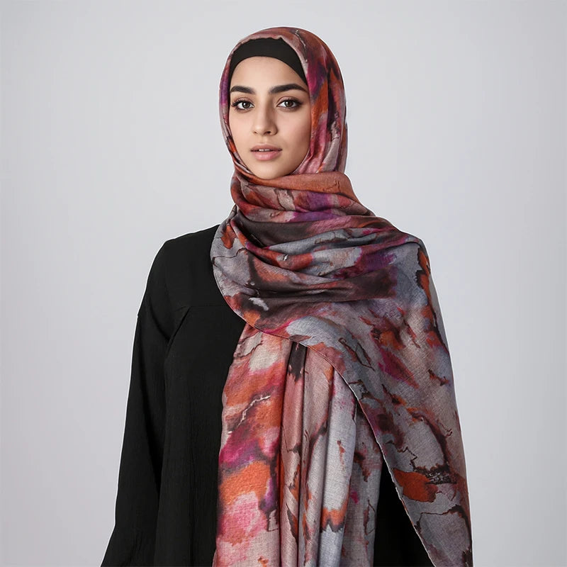 Eine Frau trägt einen fließenden Hijab-Schal mit Aquarellmuster in Orange, Pink und Grau über einem schwarzen Oberteil.