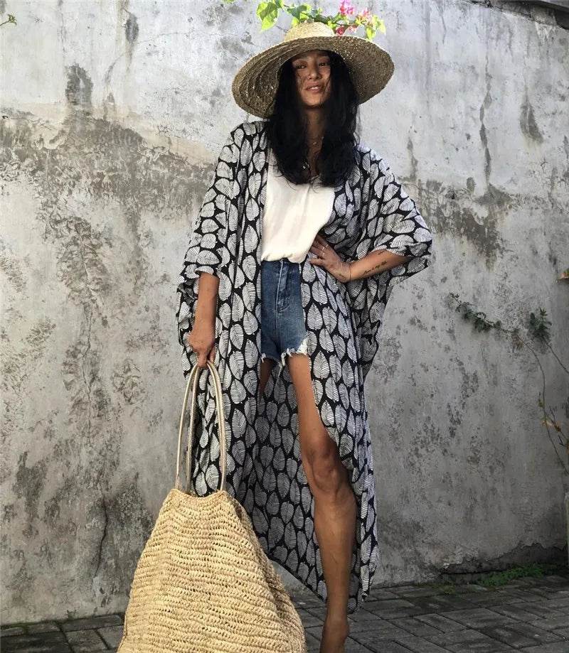 Eine Frau in einem schwarz-weißen gemusterten Kimono-Überkleid mit Jeansshorts und Strohhut vor grauem Hintergrund