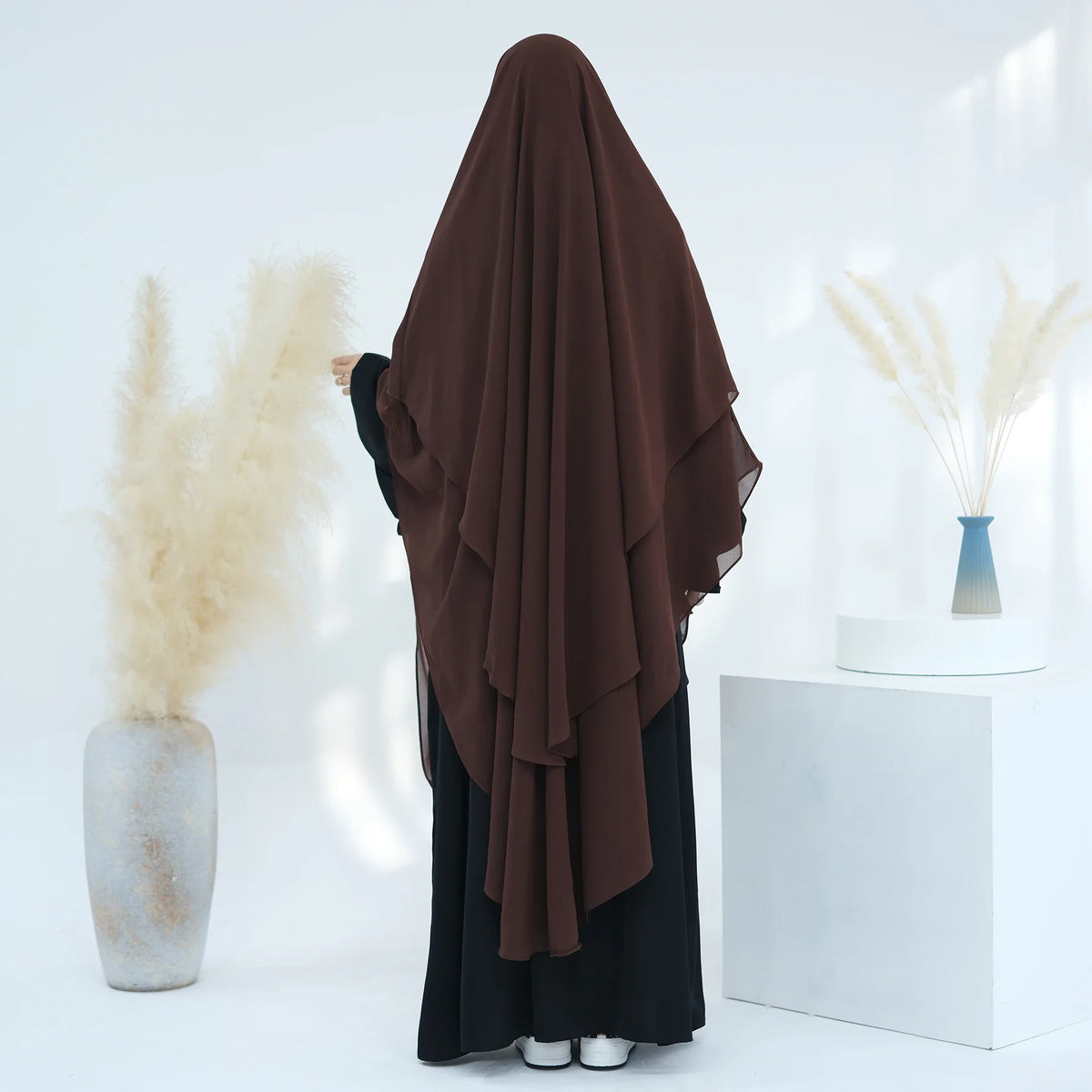 Dunkelbrauner Niqab-Kopftuch über einem schwarzen Kleidungsstück neben getrocknetem Ziergräsern.