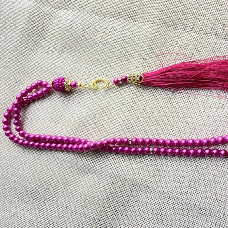 Pinkes Perlenarmband mit goldfarbener Kette und magentafarbenem Quaste auf hellem Stoff.