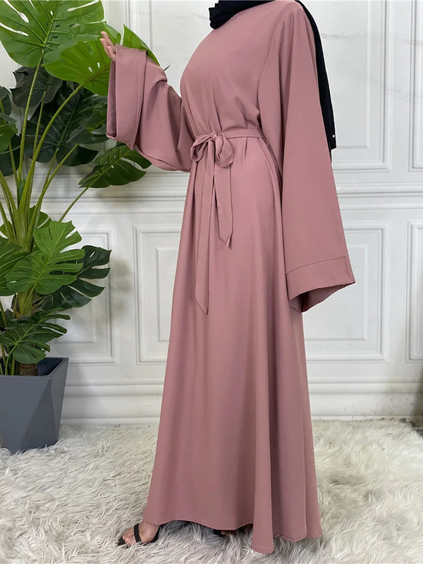 A woman wearing a dusty rose maxi abaya with black trim, standing in front of a white paneled wall.