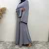 Eine Frau in einem eleganten, bodenlangen grauen Maxi-Abaya mit Ärmeln und Hijab steht vor einer weißen Wand.