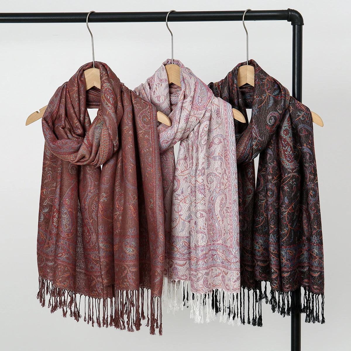 Exhibición de bufandas y chalecos bordados paisley vintage en tonos marrón, rosa y granate colgados en una barra negra.