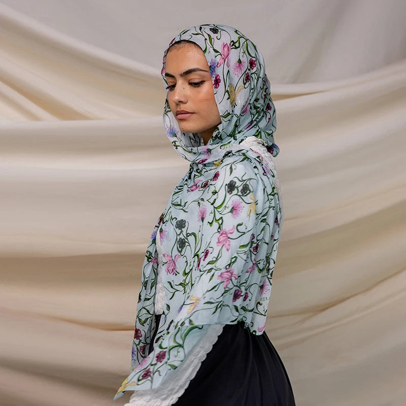 A woman wearing a light blue watercolor printed scarf hijab with floral patterns against a cream background.