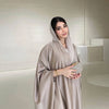 Una mujer vistiendo una abaya de satén beige con mangas de murciélago y capucha, de pie en un entorno interior luminoso.