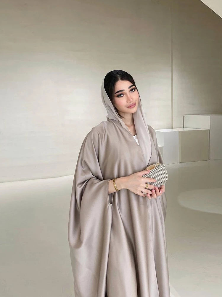 Una mujer vistiendo una abaya de satén beige con mangas de murciélago y capucha, de pie en un entorno interior luminoso.