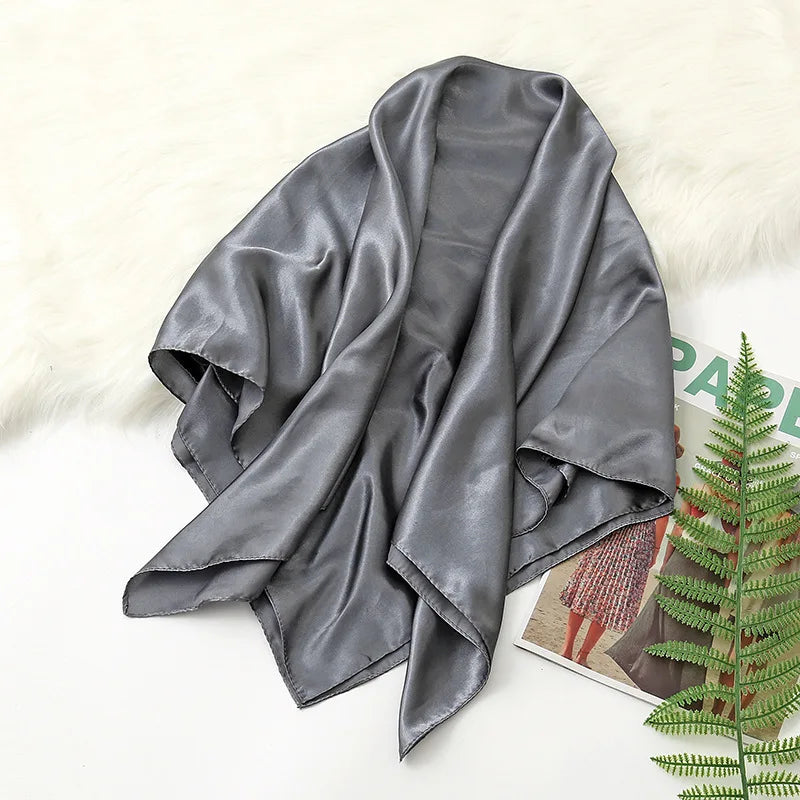 A draped, solid gray silk satin hijab scarf lying on a white surface next to a green fern.