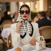 A woman wearing a white blazer and a black polka dot scarf sits at a table with a cup of coffee.