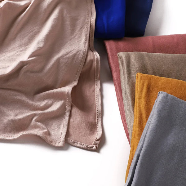 A collection of folded stretchy premium cotton hijab scarves in various muted tones including beige, dusty rose, olive green,
