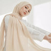Eine Frau trägt einen beigen Chiffon-Hijabschal und ein weißes Oberteil und blickt in die Kamera.
