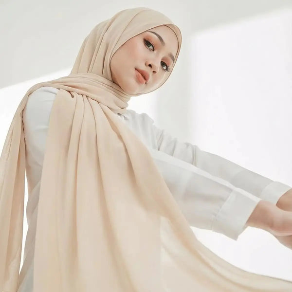 A woman wearing a beige chiffon hijab scarf and white top, looking towards the camera.