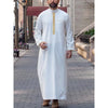 A man wearing a white embroidered long-sleeve thobe or jubbah in an outdoor setting.