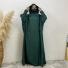 Une personne portant une abaya vert foncé avec un capuchon et des manches batwing se tient contre un mur blanc.