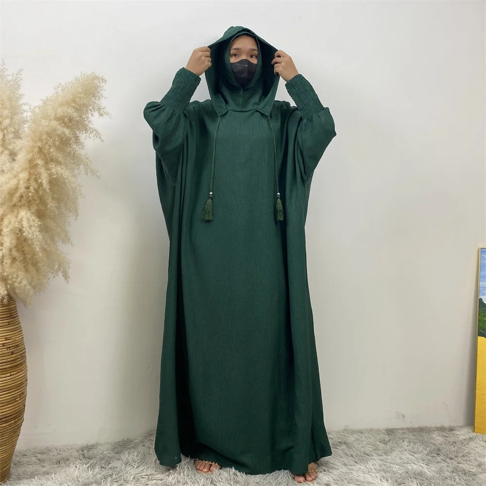 Une personne portant une abaya vert foncé avec un capuchon et des manches batwing se tient contre un mur blanc.