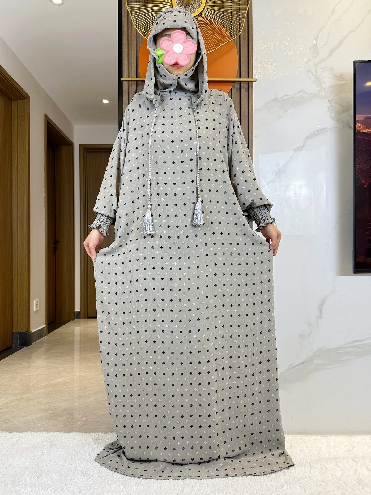 Una mujer vistiendo una abaya de algodón gris con puntos y capucha y detalle floral, de pie en interiores.