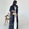 Une femme portant un trench-coat à manches longues à plissé bleu foncé par-dessus une robe blanche.
