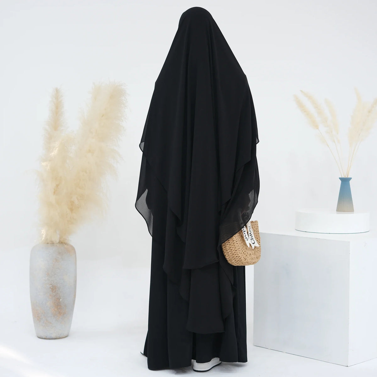 Schwarzer, zweilagiger Chiffon-Niqab-Kopftuch neben getrocknetem Ziergras und einem weißen Sockel.