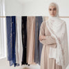 A woman wearing a beige dress and a white hijab with floral embroidery, standing in front of a rack of chiffon shawls.